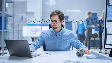 Modern Factory: Industrial Engineer Sitting at His Desk in Workshop, Working on Computer, Redrawing Blueprints, Analyzing Industrial Mechanism. Background Functional Manufactory with CNC Machinery