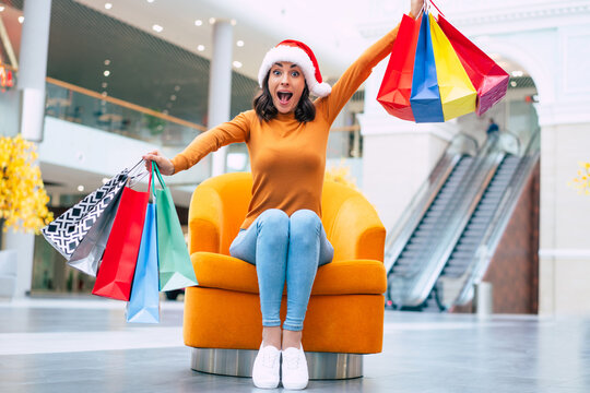 Excited Happy Beautiful Young Woman In Santa Hat With Many Colorful Bright Shopping Bags Is Sitting On The Chair In The Mall During Christmas Holiday