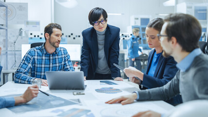 Modern Factory Office Meeting Room: Multi-Ethnic and Diverse Team of Engineers Managers Investors Talking Sitting at Table Analyzing Blueprints, Mechanism Component. High-Tech Manufactory Optimization