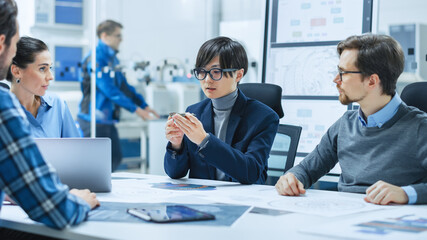 Factory Office Meeting Room: Computer Design Specialist Holds Printed Circuit Board Prototype and Reports to Diverse Team of Engineers, Managers Sitting at Conference Table. Industrial Facility