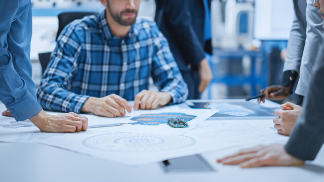 Busy Diverse Team of Computer Engineers and Specialists Gather Around Conference Table and Discuss Electrical Prototype, Drafts and Blueprints, Find Problem Solutions. Office Meeting Room.