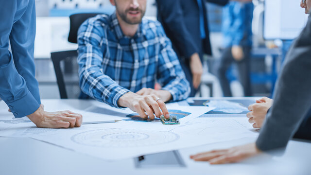 Busy Diverse Team of Computer Engineers and Specialists Gather Around Conference Table and Discuss Electrical Prototype, Drafts and Blueprints, Find Problem Solutions. Office Meeting Room.