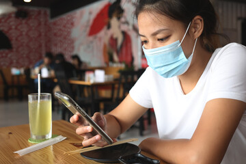 Asian woman wearing a mask in a restaurant with holding the smartphone. Women wear face masks to prevent communicable diseases.