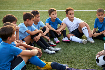 Obraz premium group of happy caucasianteen boys in football uniforms having a rest on the sport field, sportive children take a break