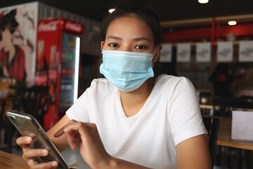 Asian woman wearing a mask in a restaurant with holding the smartphone. Women wear face masks to...