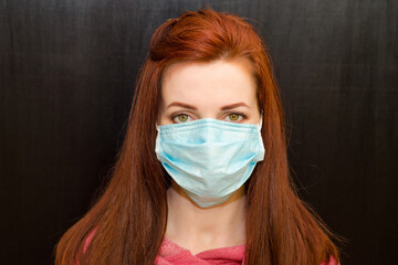 Young caucasian redhaired girl with green eyes, in medical mask, pink sweater on black background,...