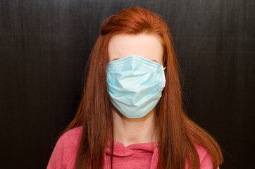 Young caucasian redhaired girl with green eyes, in wrong used medical mask, on black background,...
