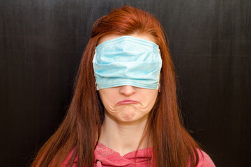 Young caucasian redhaired girl with green eyes, in wrong used medical mask, on black background,...