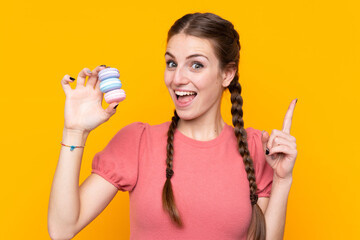 Young woman over isolated yellow background holding colorful French macarons and pointing up a great idea