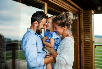 Family with small daughter standing on patio of wooden cabin, holiday in nature concept. - obrazy, fototapety, plakaty