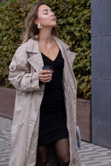 beautiful slim young brunette in fashionable trench coat and black dress walking through the...
