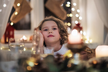 Bored small girl sitting at the table indoors at Christmas.