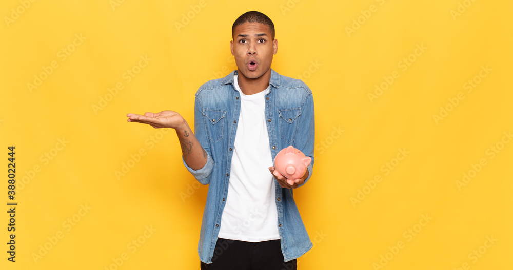 Wall mural young black man looking surprised and shocked, with jaw dropped holding an object with an open hand on the side