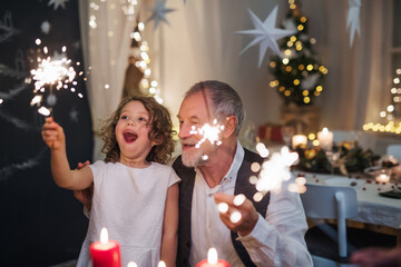 Senior grandfather with small granddaughter indoors at Christmas, sitting at table with sparklers.