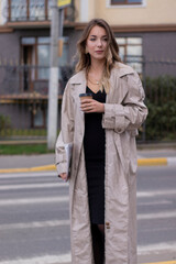 Fototapeta premium beautiful slim young brunette in fashionable trench coat and black dress cross a pedestrian crossing.