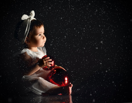 Little female in white headband and dress, barefoot. She holding two red balls, sitting on floor. Twilight, black background.