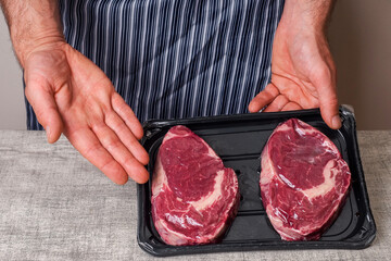 Butcher holds plastic tray with two premium rib eye steaks with high quality marbling. One hand points to the steaks. Meat industry product.