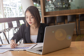 Fototapeta premium A beautiful young businesswoman sits on a desk using a laptop and checks the graph table to calculate the financial statements within her work station.