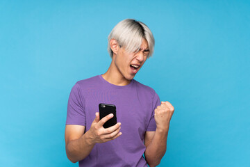 Young asian man over isolated blue background with phone in victory position