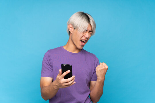 Young Asian Man Over Isolated Blue Background With Phone In Victory Position