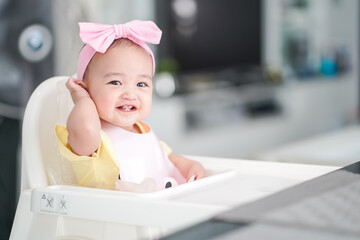 10-month old asian baby girl in a pink bib and hair bow sitting on a white high chair is happy during having a breakfast at home