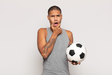 young black man with mouth and eyes wide open and hand on chin, feeling unpleasantly shocked,...