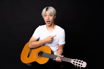 Young asian man with guitar over black background surprised and pointing side