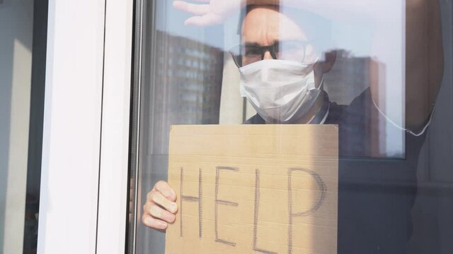 Sick white man wear black glasses, white medical disposable face mask, leaned against window. Young person with sad tired look isolated at home. Guy hold sign Help. Lock down concept during quarantine