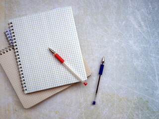 Open notebooks are stacked. The checkered pages of the notebook are clean, without notes. There are red and blue pens on the notebook and next to it. Empty space.