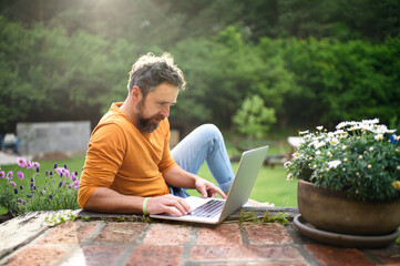 Mature man with laptop working outdoors in garden, home office concept.