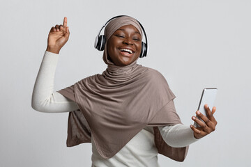 Joyful black muslim woman in hijab and headphones listening music on smartphone
