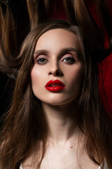 A beautiful girl with evening make-up and red lips wearing a blouse with a deep neckline and her hair suspended on chains, posing on a red and black background. Conceptual, avant-garde design