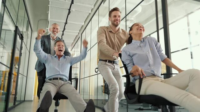 Joyful business team celebrating success. Four crazy happy colleagues riding chairs in office corridor, laughing, shouting and having fun