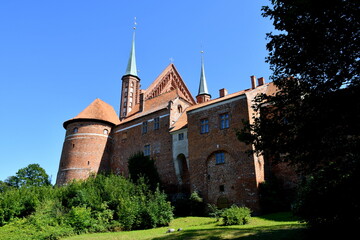 Fototapeta premium View of an old medieval castle made out of red brick with many towers, walls, and the top being a remnant of a church seen next to a public park on a cloudless summer day on a Polish countryside