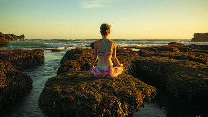 Woman sitting on the rock, practicing yoga and enjoying ocean view. Lotus pose. View from back....