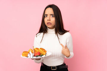 Young brunette woman holding waffles over isolated pink background pointing to oneself