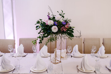 Table covered with a tablecloth and served with dishes and cutlery. Festive table decorated with composition of flowers and greenery in the wedding banquet hall.