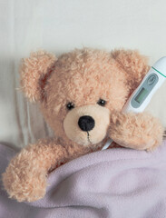 Fever, cold, flu. Cute teddy with a thermometer in bed, covered with a warm blanket