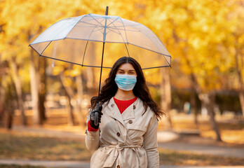 Pretty millenial lady with umbrella and medical mask standing under rain and looking at camera at autumn park