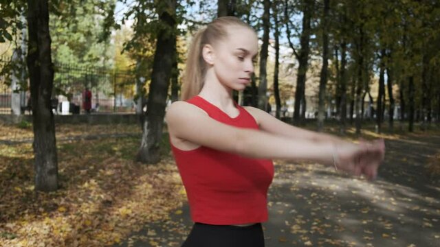 young caucasian busty woman stretching neck and arms outdoors, running away. pretty female in sportswear working out at autumn park. transport and unrecognisable people out of focus in background