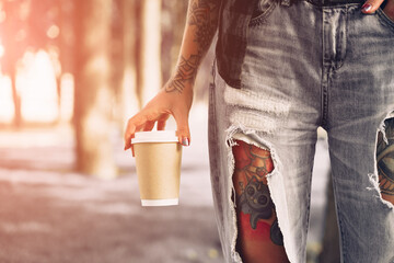 Tattooed hipster girl in torn jeans with a cup of coffee or tea. standing in the park