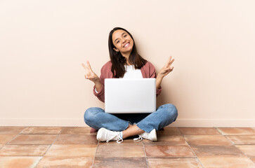 Young mixed race woman with a laptop sitting on the floor showing victory sign with both hands