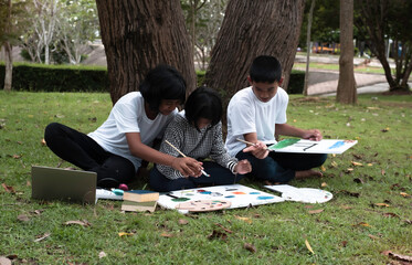 Three children sitting on green grass ground floor,painting color on canvas and talking,doing activity together with happy feeling,in a park