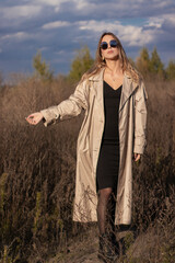 brunette in fashionable trench coat, black dress and sunglasses in the field. autumn, yellow leaves, blue sky.