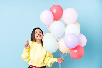 Woman holding balloons in a party over isolated blue background with fingers crossing
