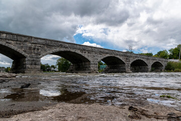 The historic Pakenham five-arch stone bridge in Pakenham, Ontario reflects in the river beneath it on a mostly cloudy day.