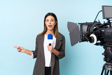 Reporter woman holding a microphone and reporting news over isolated blue background surprised and pointing finger to the side