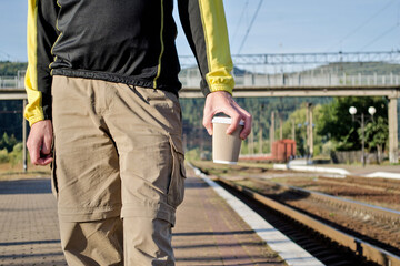 Young man in sport tourist clothes stay on the platform of the station, near the rails, and holds a cup of coffee