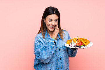 Brunette woman holding waffles over isolated pink background whispering something