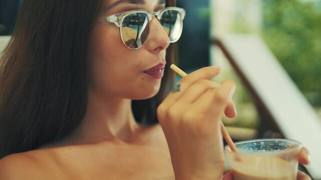 young girl is sunbathing by the pool and sipping a drink from a straw. in sunglasses and brown hair.